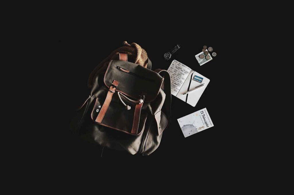 Travel essentials laid out on a black background, featuring a sturdy backpack, a notebook with travel plans, a pen, some local currency, a wristwatch, and a book, showcasing the must-haves for an organized and prepared journey.