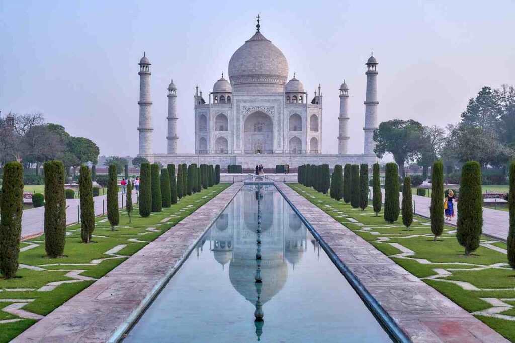 The Taj Mahal with its iconic white marble dome and minarets, reflected in a long rectangular pool, surrounded by lush gardens and visitors.