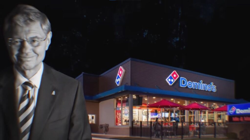 A man in a suit stands in front of a Domino's Pizza restaurant at night.