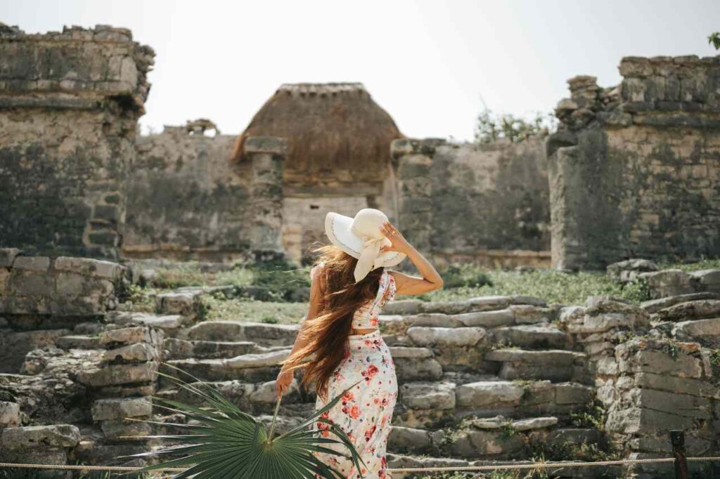 Woman in a floral dress with a wide-brimmed hat standing in front of ancient ruins in Tulum, Mexico.